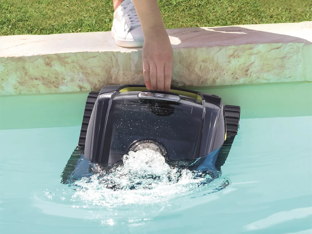 A hand lowering the cleaner into a pool off the coping, water churning around it